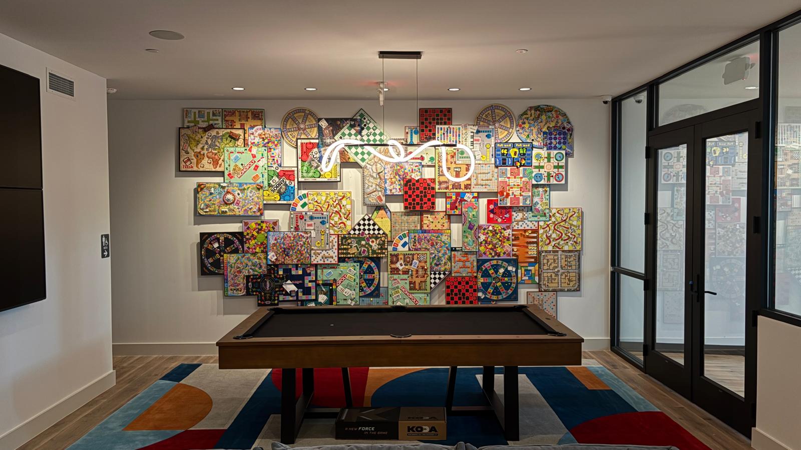 Wall collage of vintage board games behind a pool table with a neon light fixture, in an amenity lounge