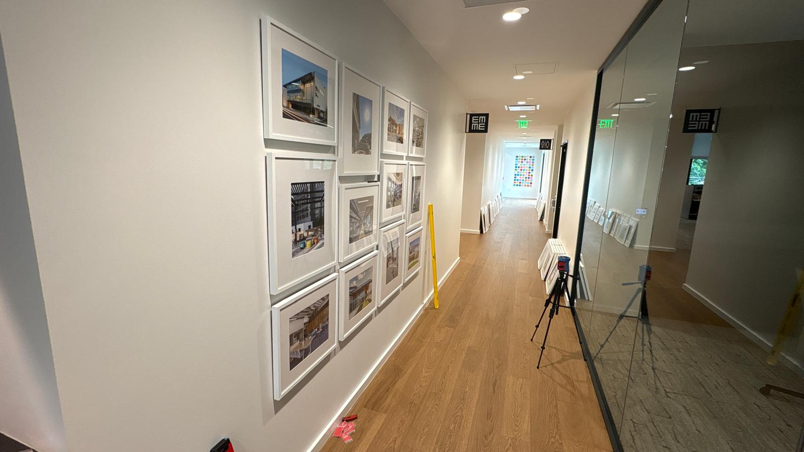 Twelve framed architectural photographs being installed along a hallway wall in an office space