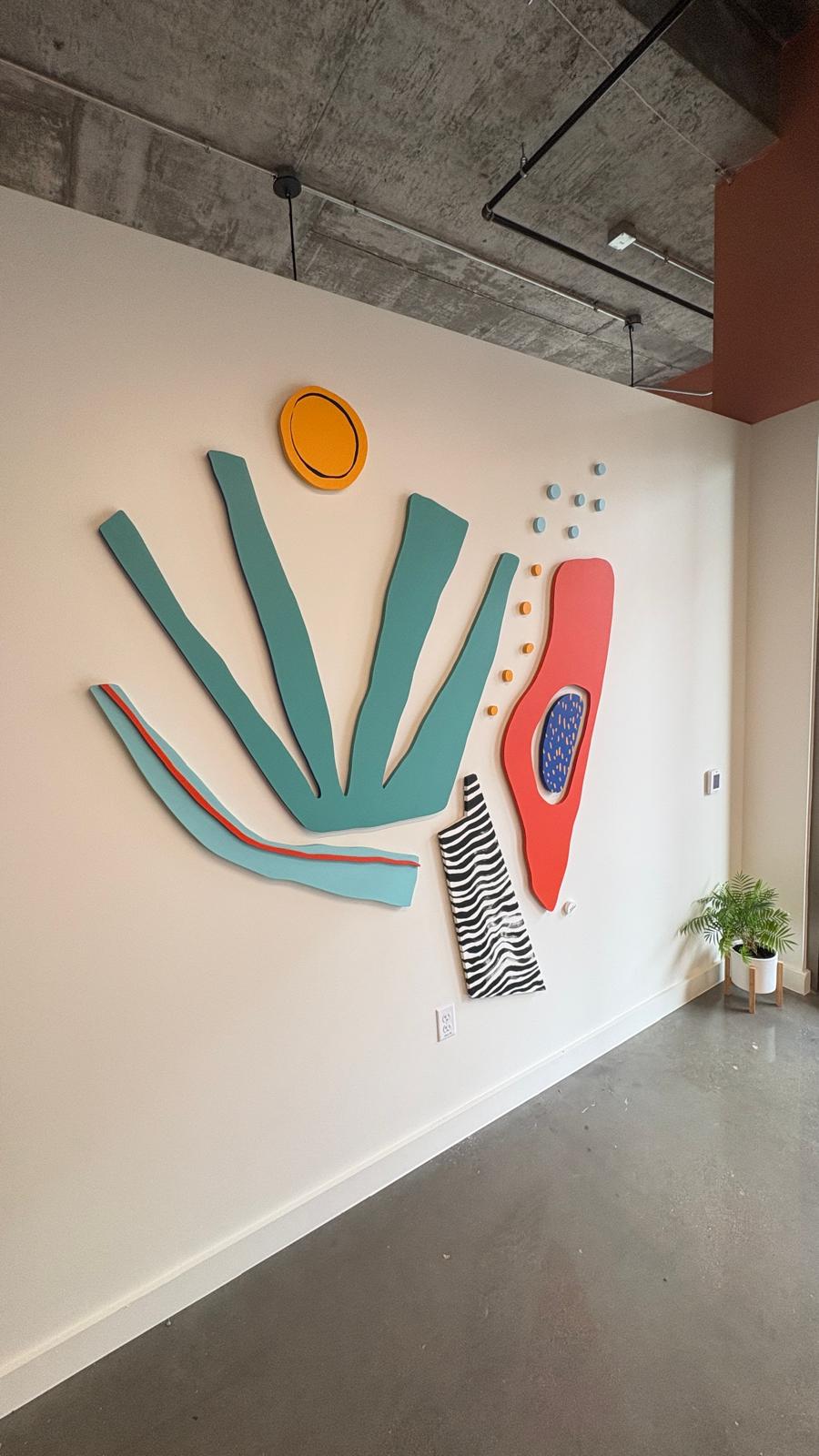 Abstract sculptural wall art with organic shapes in teal, orange, and red on a concrete-ceilinged commercial wall